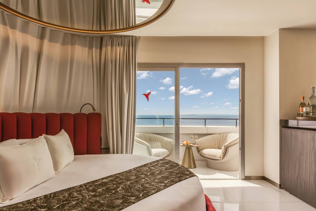Ein Hotelzimmer mit Bett, einem Balkon mit Meerblick, einem Essbereich und einer Minibar.
