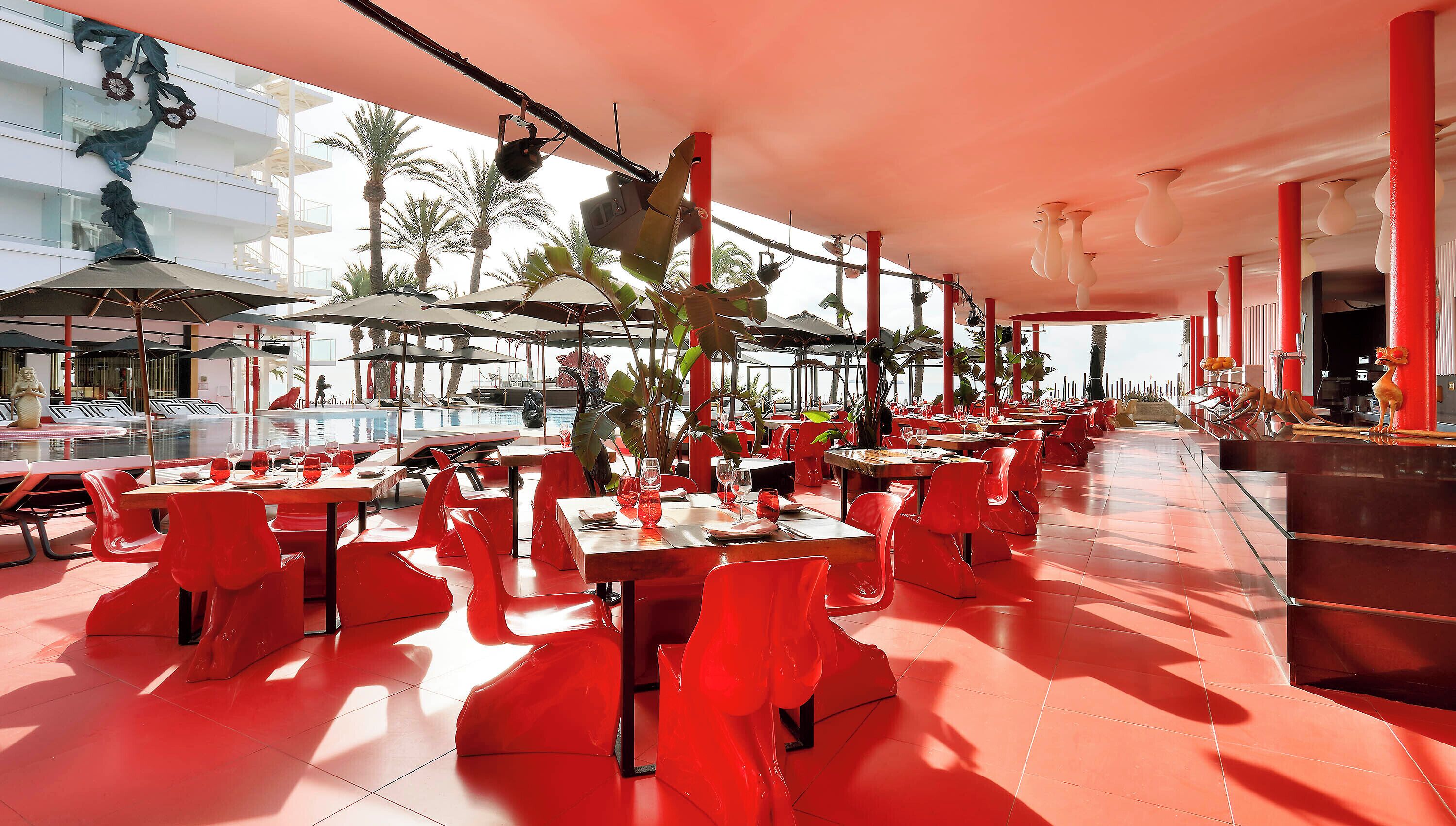 Un amplio espacio exterior para comer con sillas y mesas rojas, rodeado de sombrillas y palmeras.