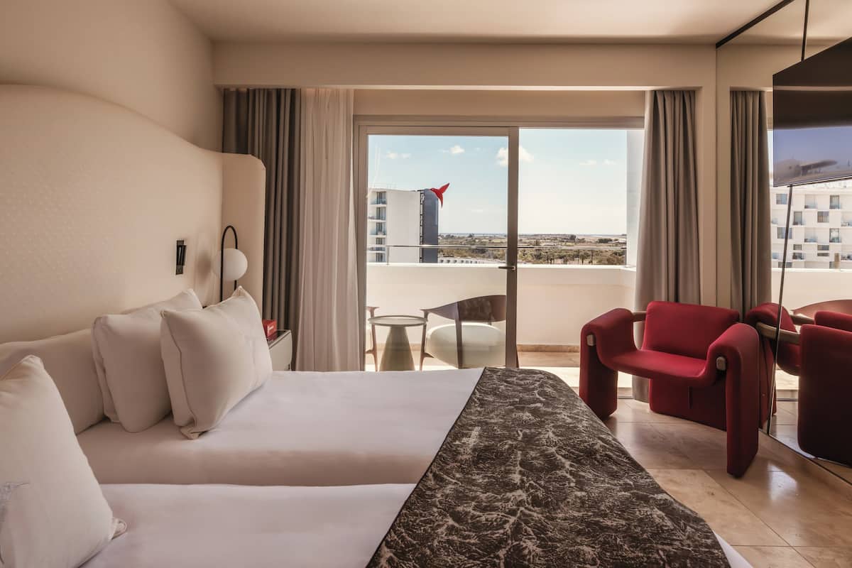 Ein Hotelzimmer mit Bett, einem roten Sessel, einem kleinen Tisch und Blick auf die Stadt durch große Fenster.