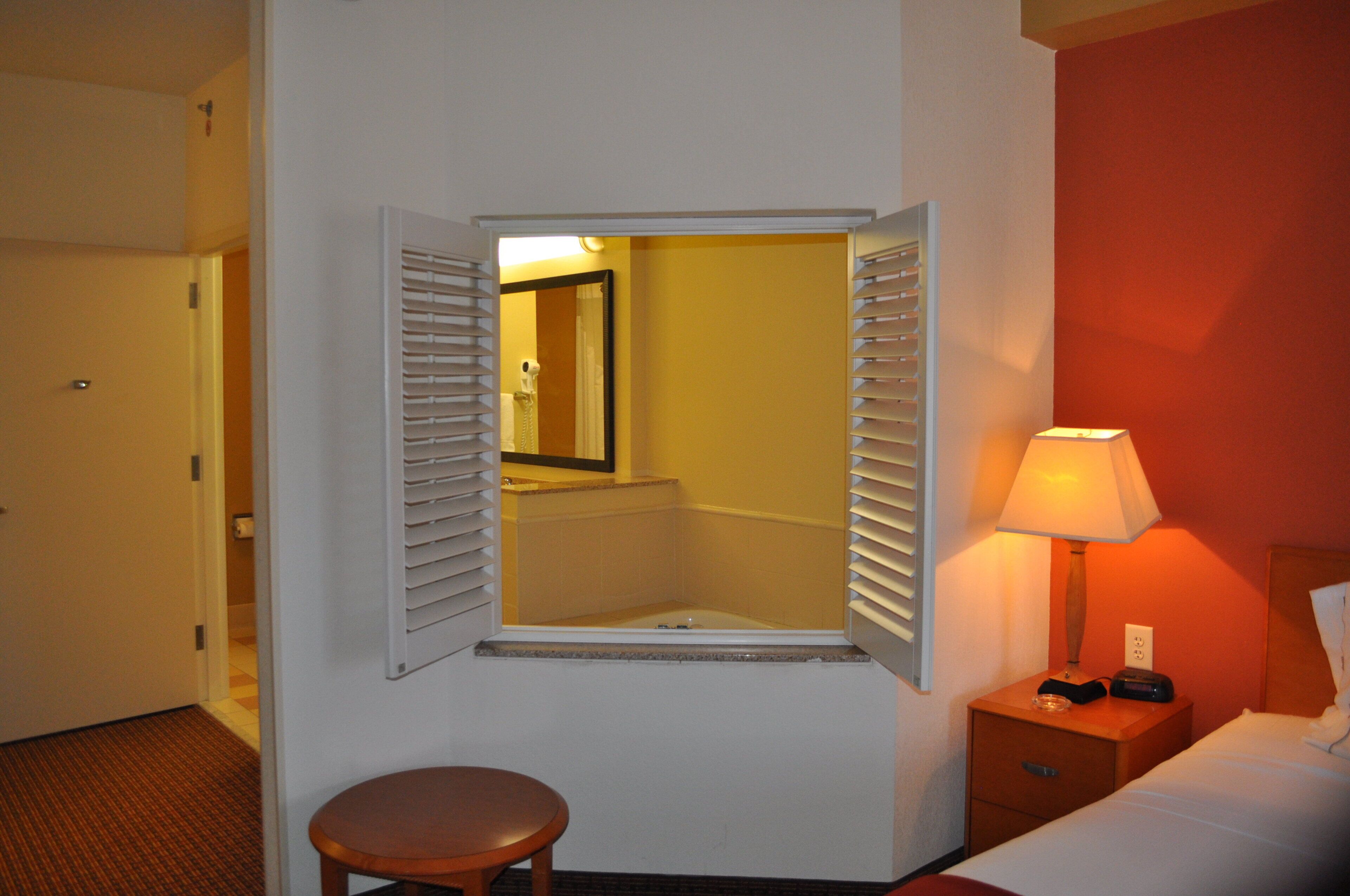 A hotel room with a single bed, a bedside table with a lamp, a small wooden stool, and a window with blinds.