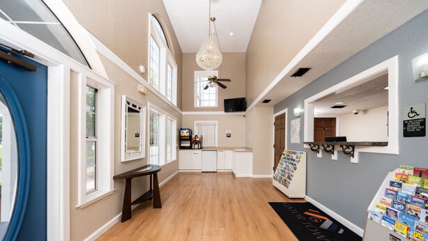 A hotel lobby with a wooden floor, a chandelier, a ceiling fan, a reception desk, and a wall-mounted information board. - Gator Town Inn (Gainesville)