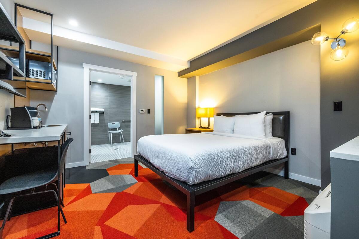 A modern hotel room with a bed, desk, chair, and a geometric patterned carpet.