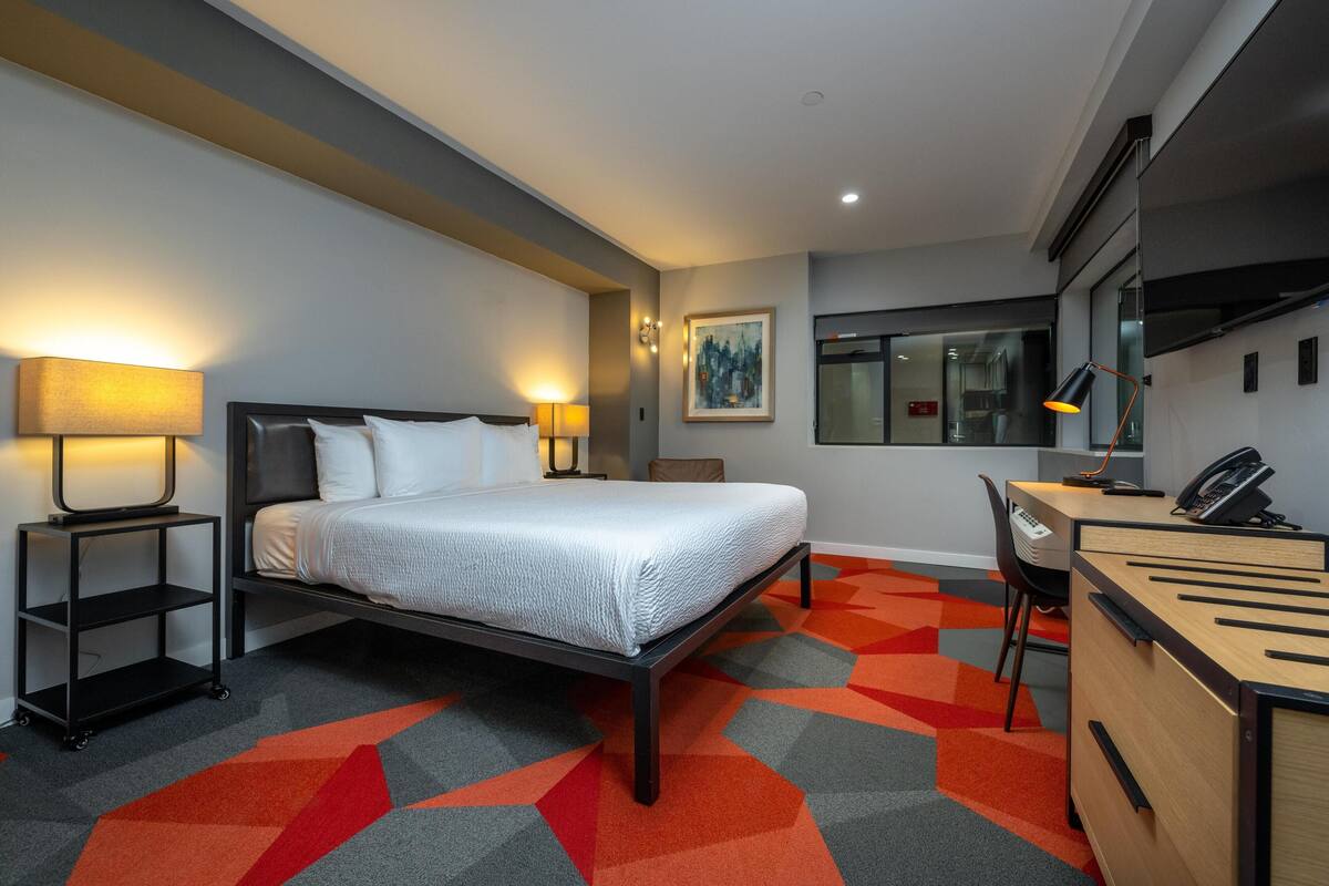 A hotel room with a bed, desk, chair, lamp, and a painting on the wall.