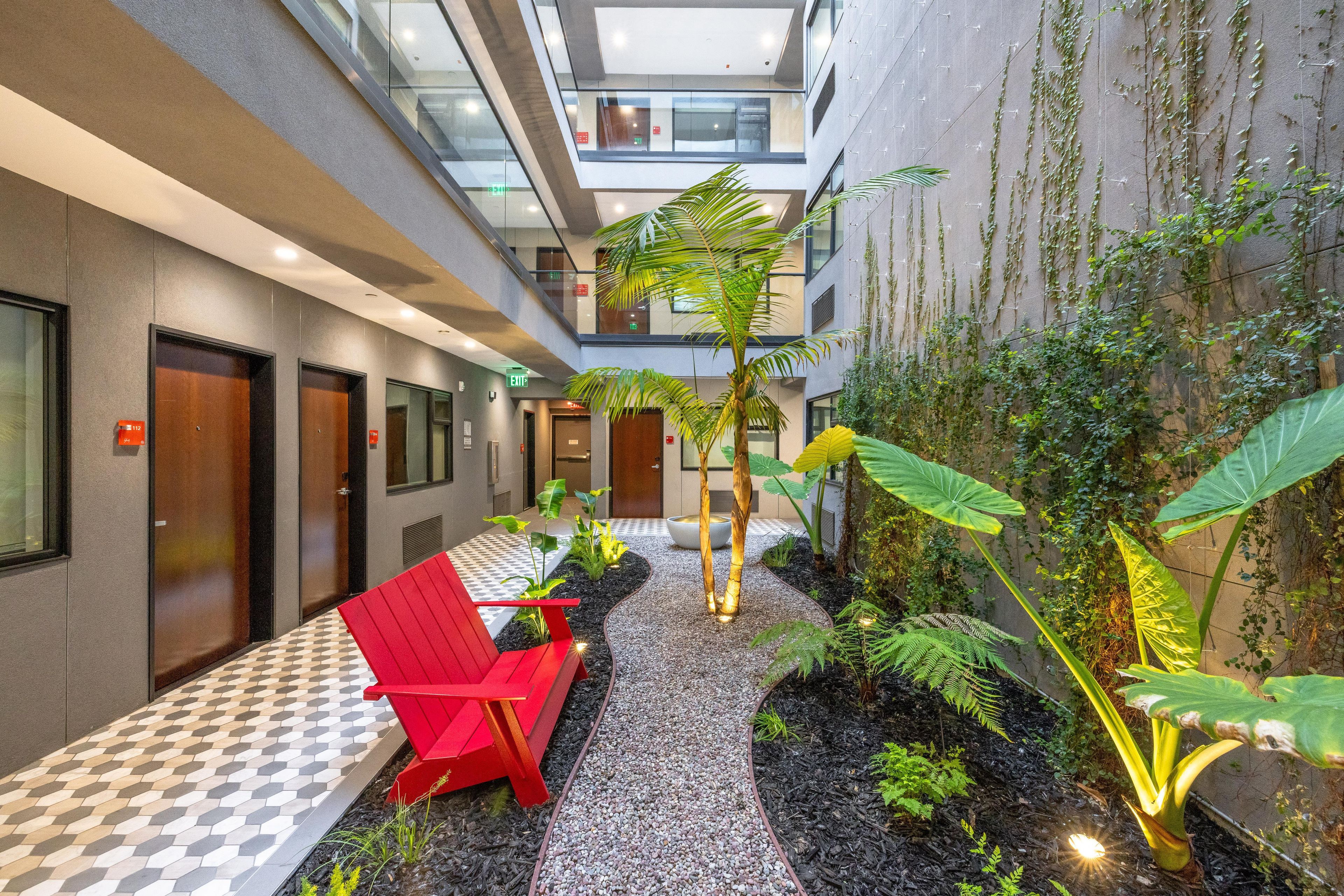Un couloir d’hôtel avec une chaise rouge, un palmier et un mur recouvert de plantes grimpantes.