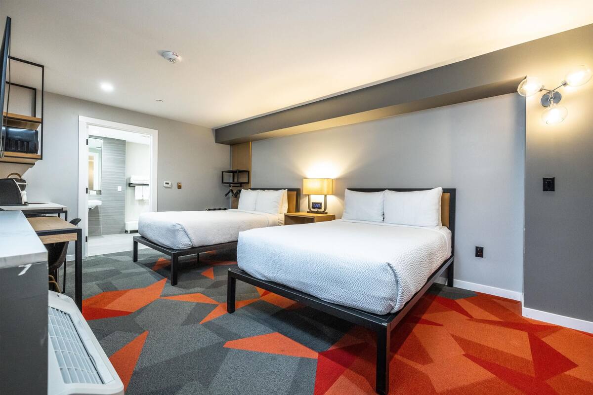 A hotel room with two beds, a desk, a chair, a TV, and a bathroom visible through an open door.