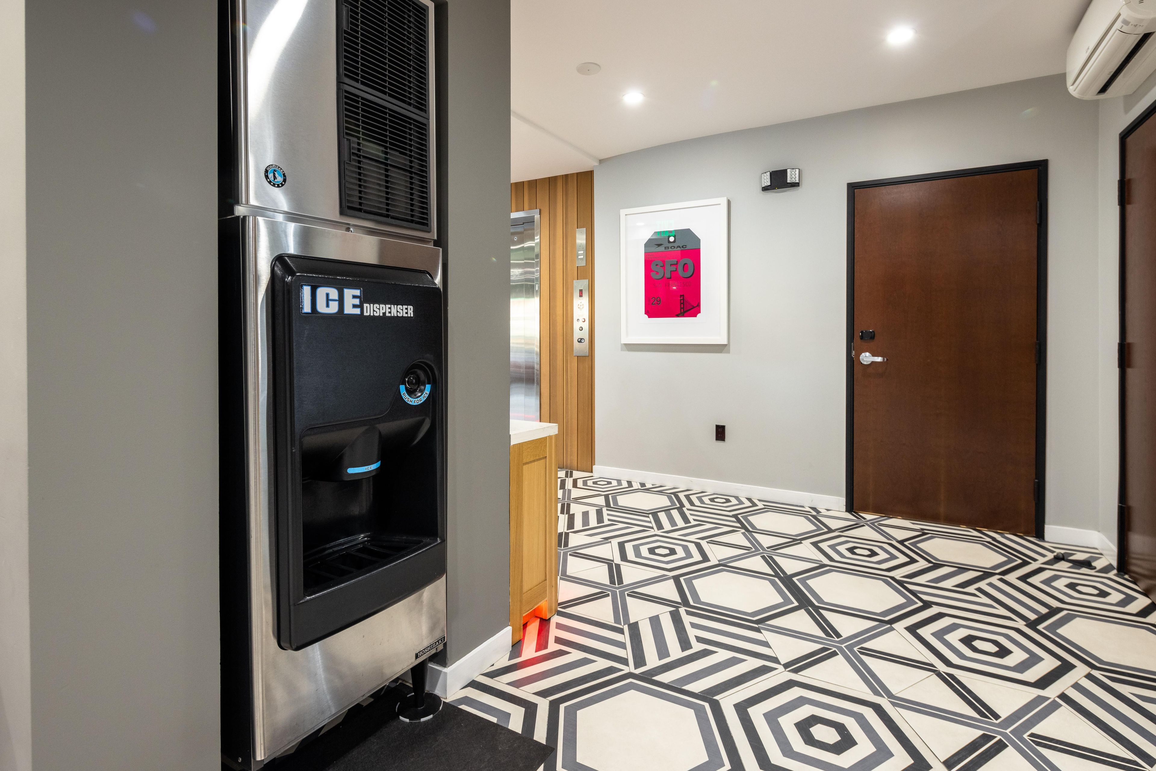 Un couloir d’hôtel avec un distributeur de glaçons moderne, une œuvre d’art encadrée au mur et un sol à motifs.