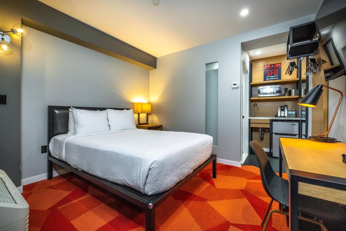 A hotel room with a bed, a desk, a chair, a lamp, a microwave, and a small kitchenette.
