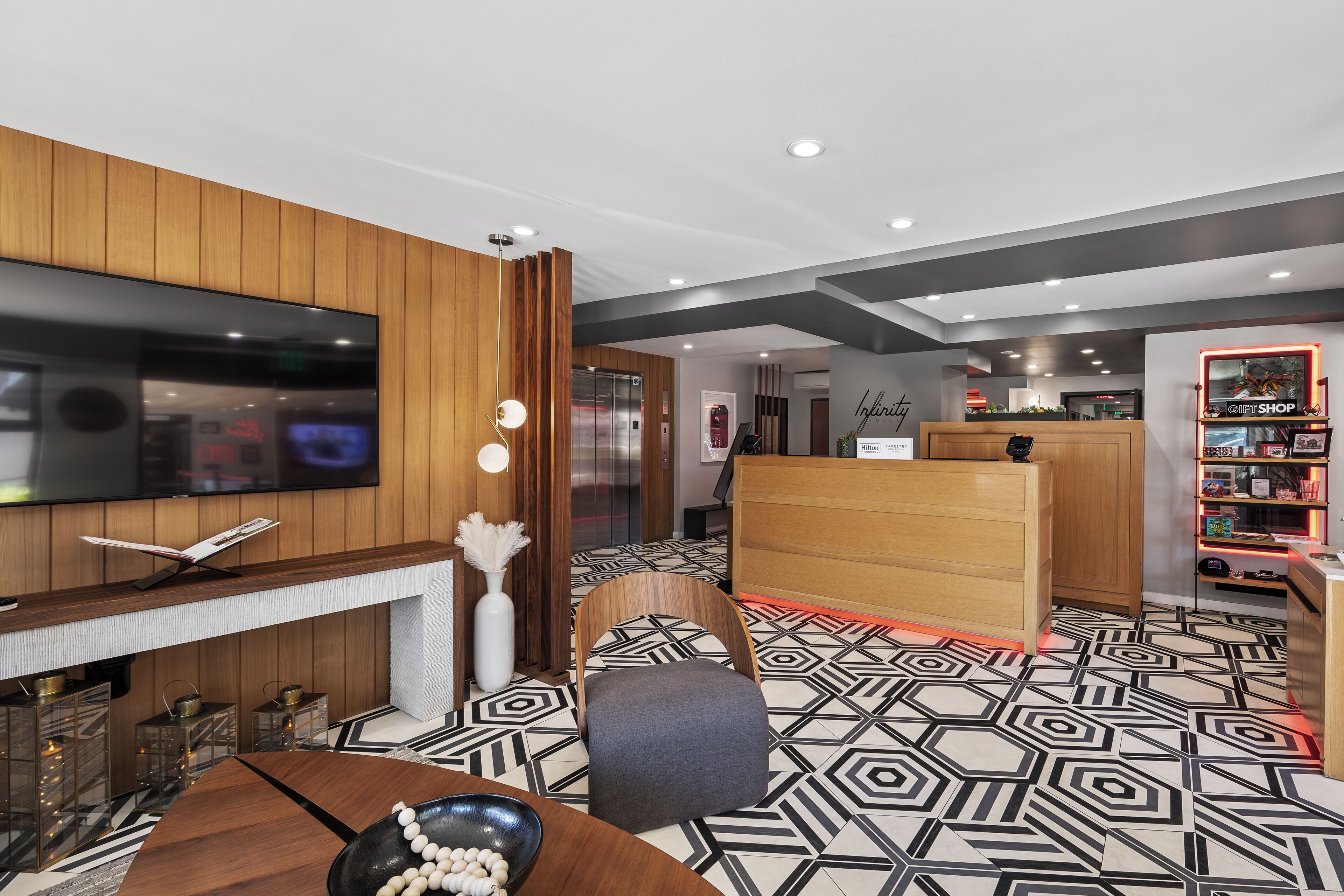 A hotel lobby with a modern design, featuring a flat-screen TV, a fireplace, a reception desk, and a patterned floor.