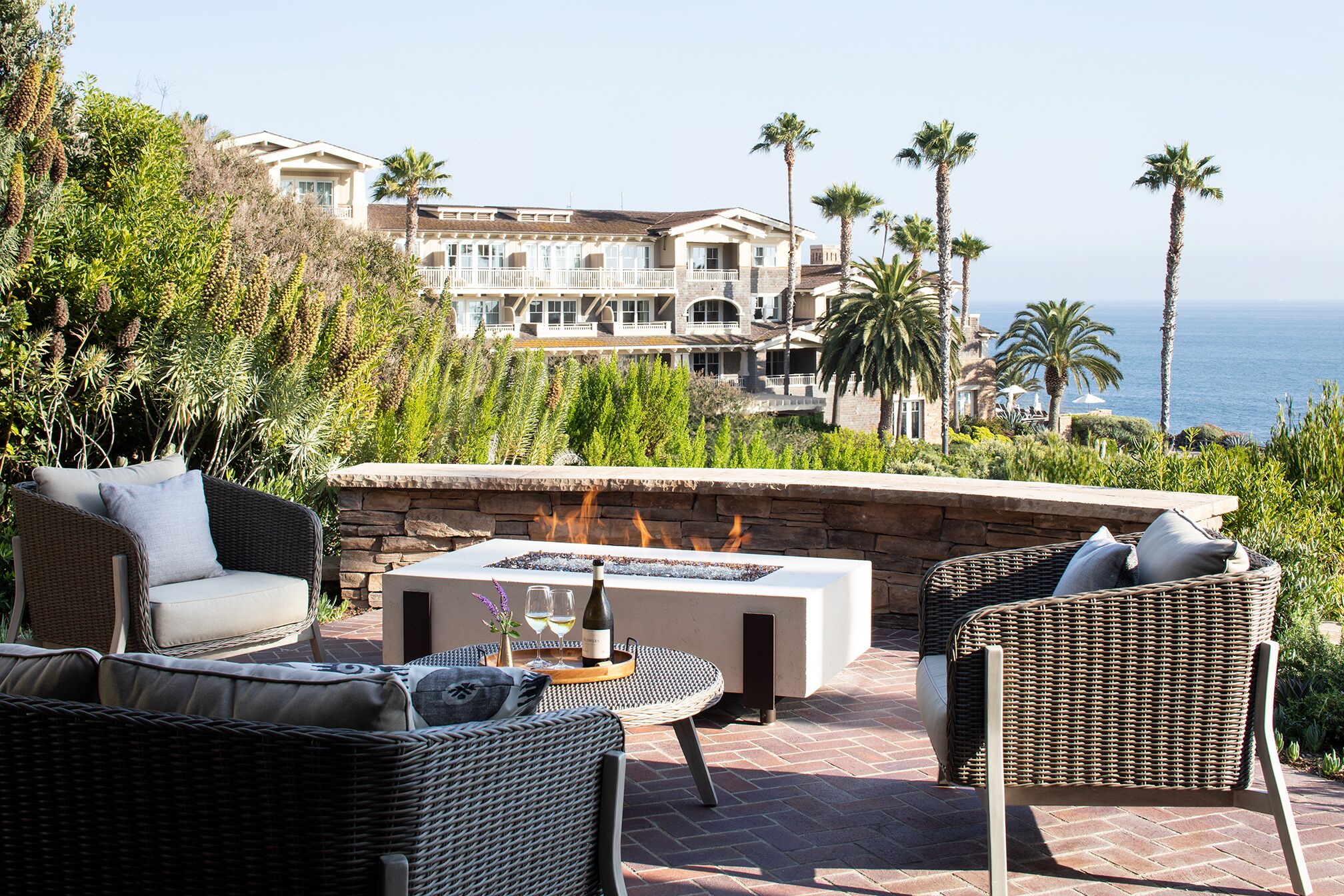 A patio with wicker furniture, a stone fire pit, and a view of a coastal estate.