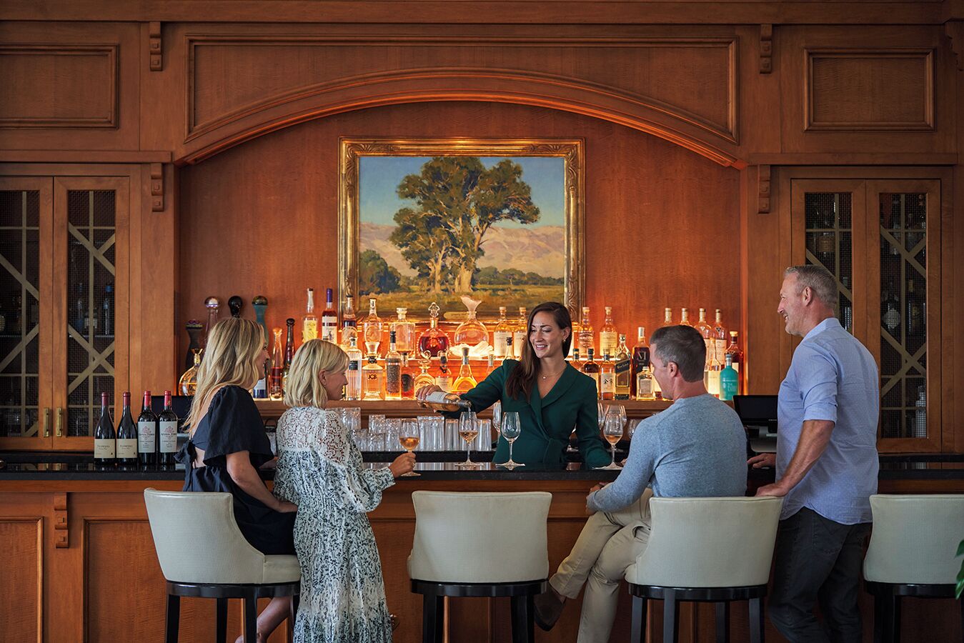 A bar area with wooden paneling, a painting of a tree, and various bottles of alcohol.
