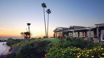 A coastal property with a large house featuring a covered patio and a view of the ocean at sunset.