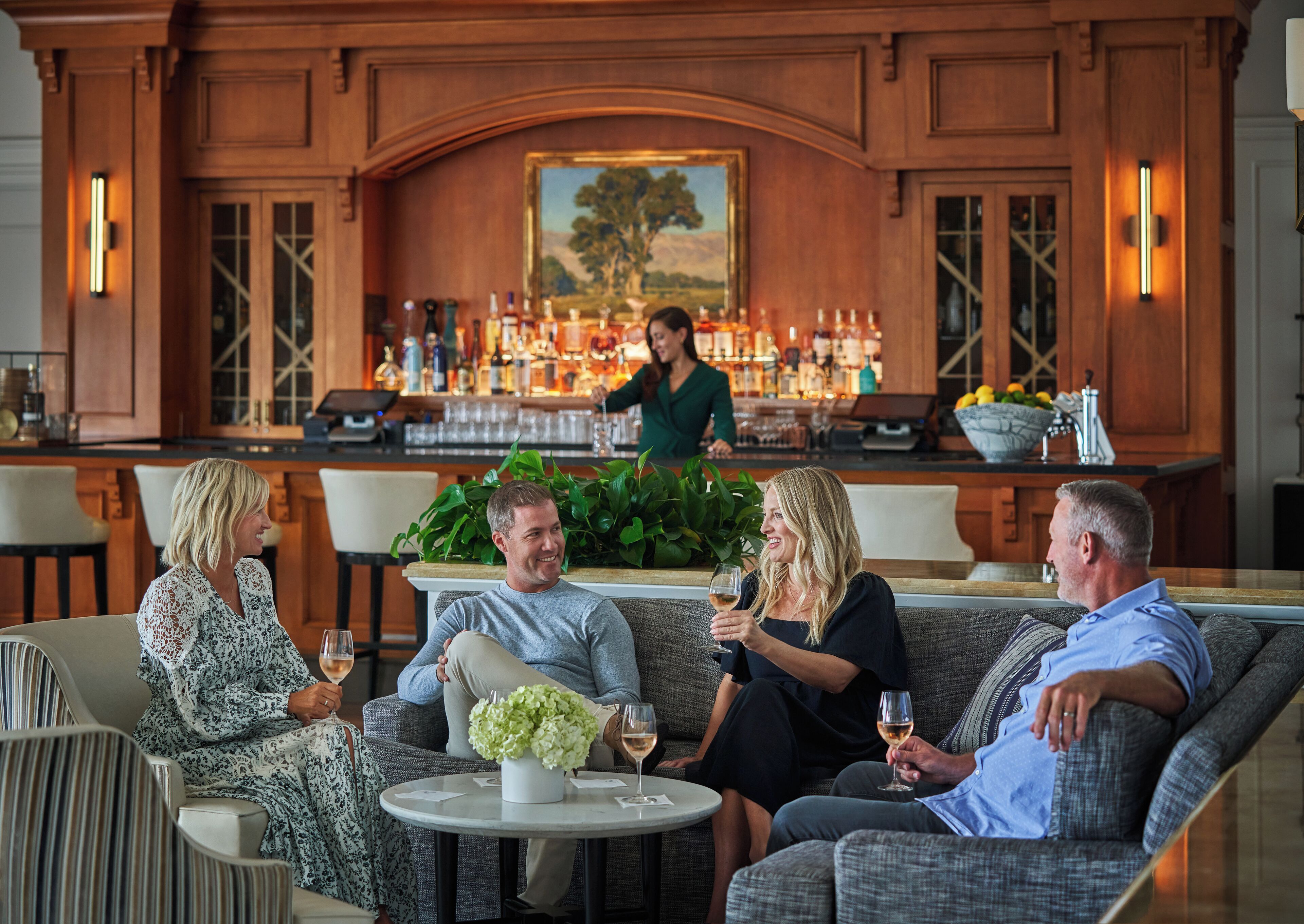 Eine Bar mit Theke, Barkeeper und einer Sitzecke mit Sofas und einem runden Tisch mit Blumen.