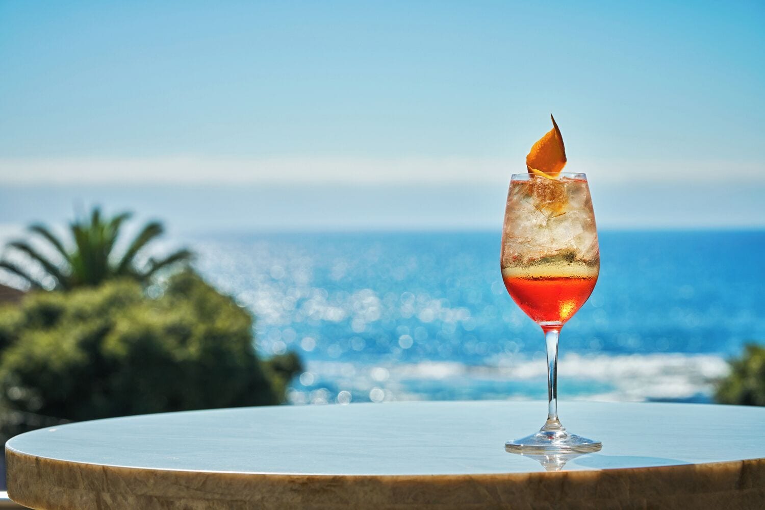A cocktail glass with a flaming garnish on a round wooden table, set against a scenic ocean view.
