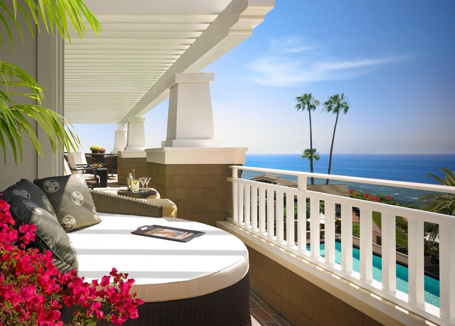 A balcony with a white railing, a wicker sofa, a small table with drinks, and a view of the ocean and palm trees.