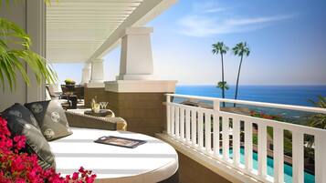 A balcony with a white railing, a wicker sofa, a small table with drinks, and a view of the ocean and palm trees.