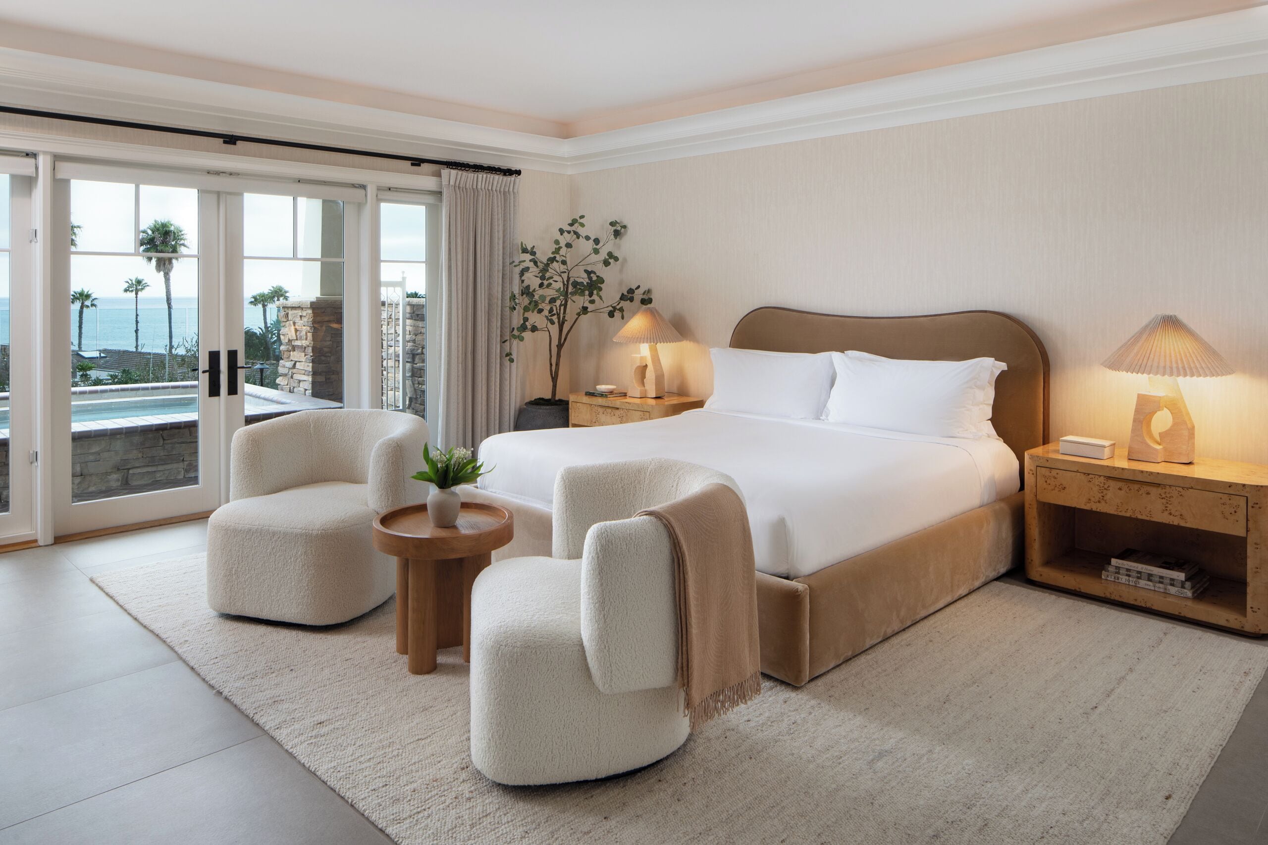 Habitación de hotel moderna con una cama grande, dos sillones, una mesita con una planta y vistas al océano a través de amplios ventanales.