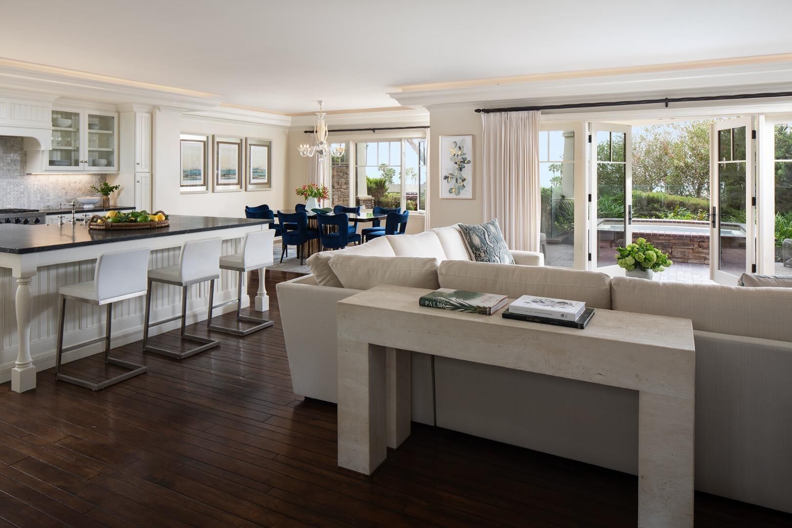 Una cocina moderna con una isla central, una zona de comedor con sillas azules, una sala de estar con un sofá grande y vistas a un jardín a través de amplios ventanales.