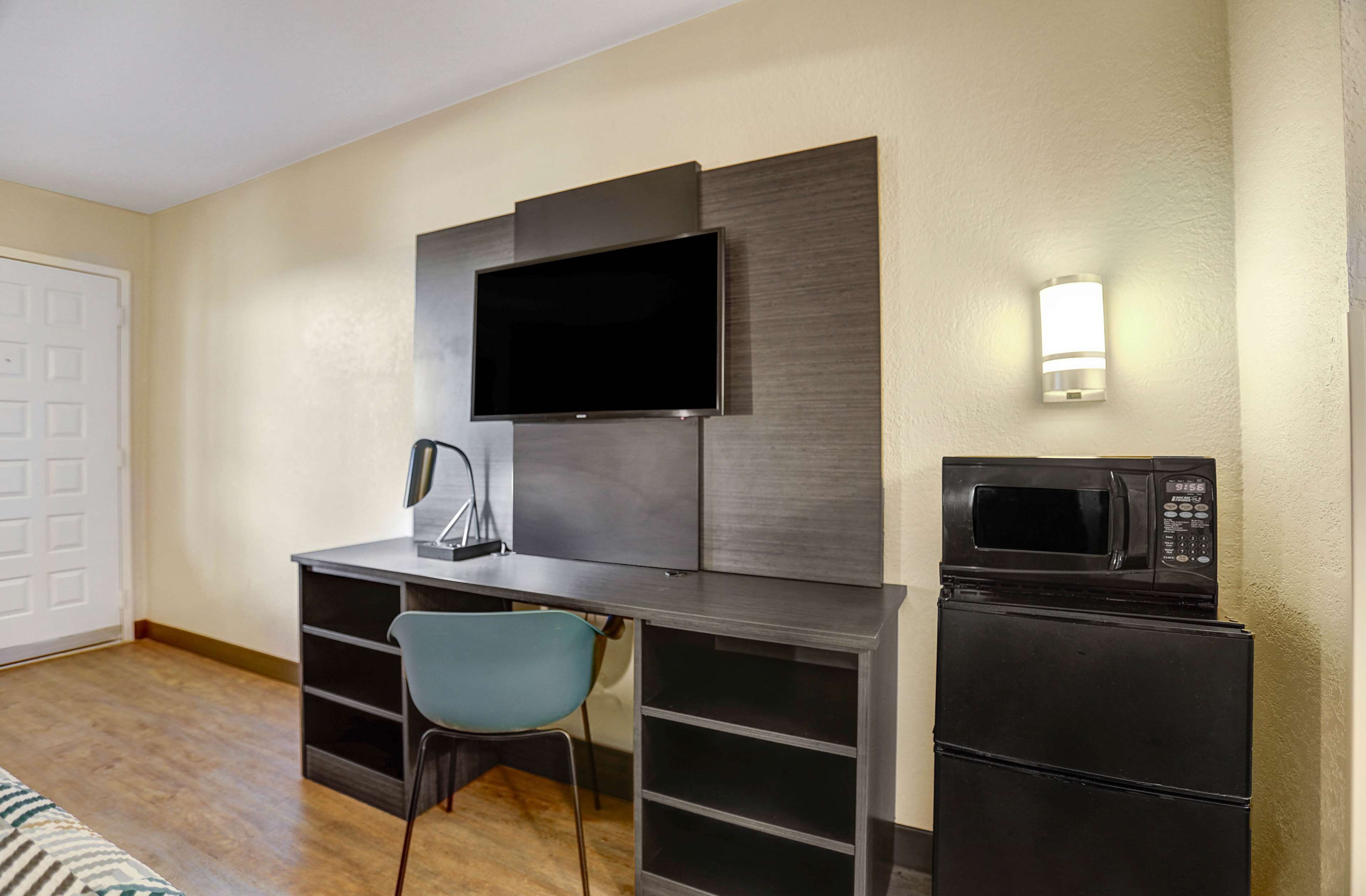 A compact room with a flat-screen TV mounted on the wall, a desk with a chair, a microwave, and a refrigerator.