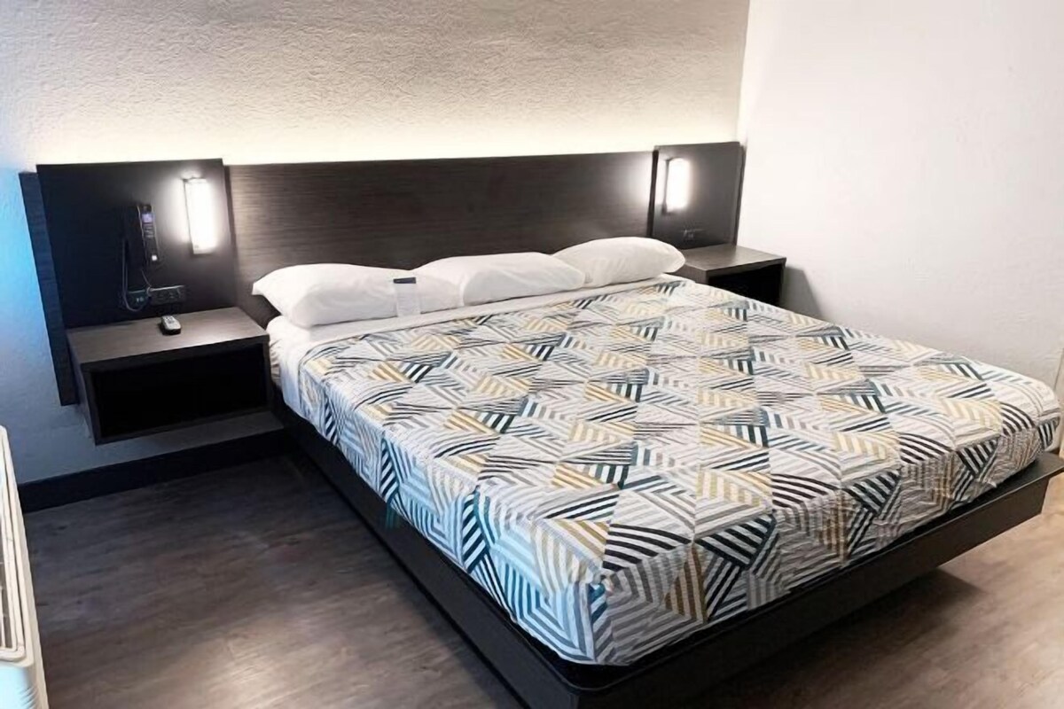 A modern bed with a geometric patterned bedspread, a dark headboard with integrated lighting, and a small bedside table with a power outlet.
