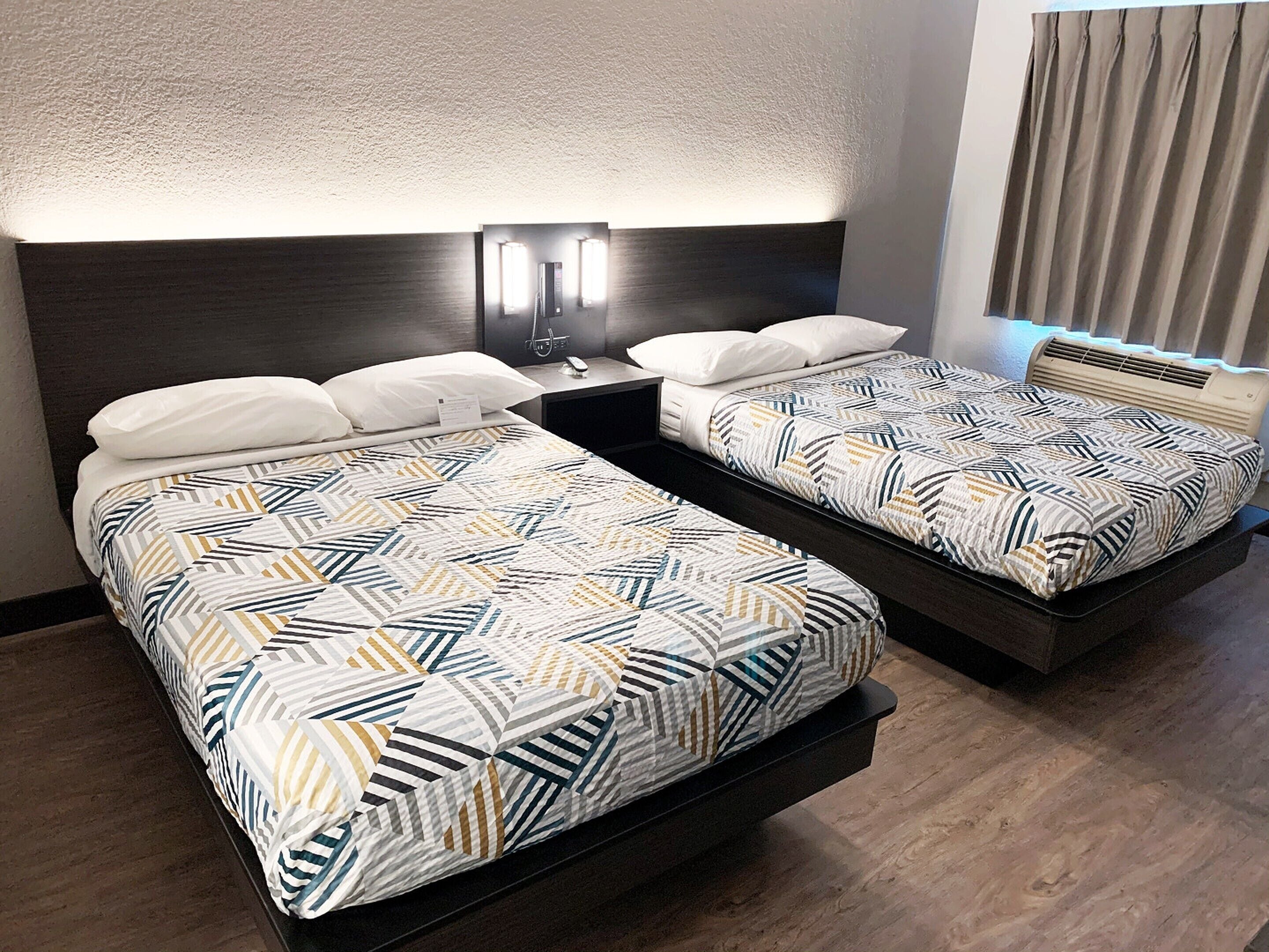 A hotel room with two beds, each with patterned bedding, a nightstand with a lamp, and a window with curtains.