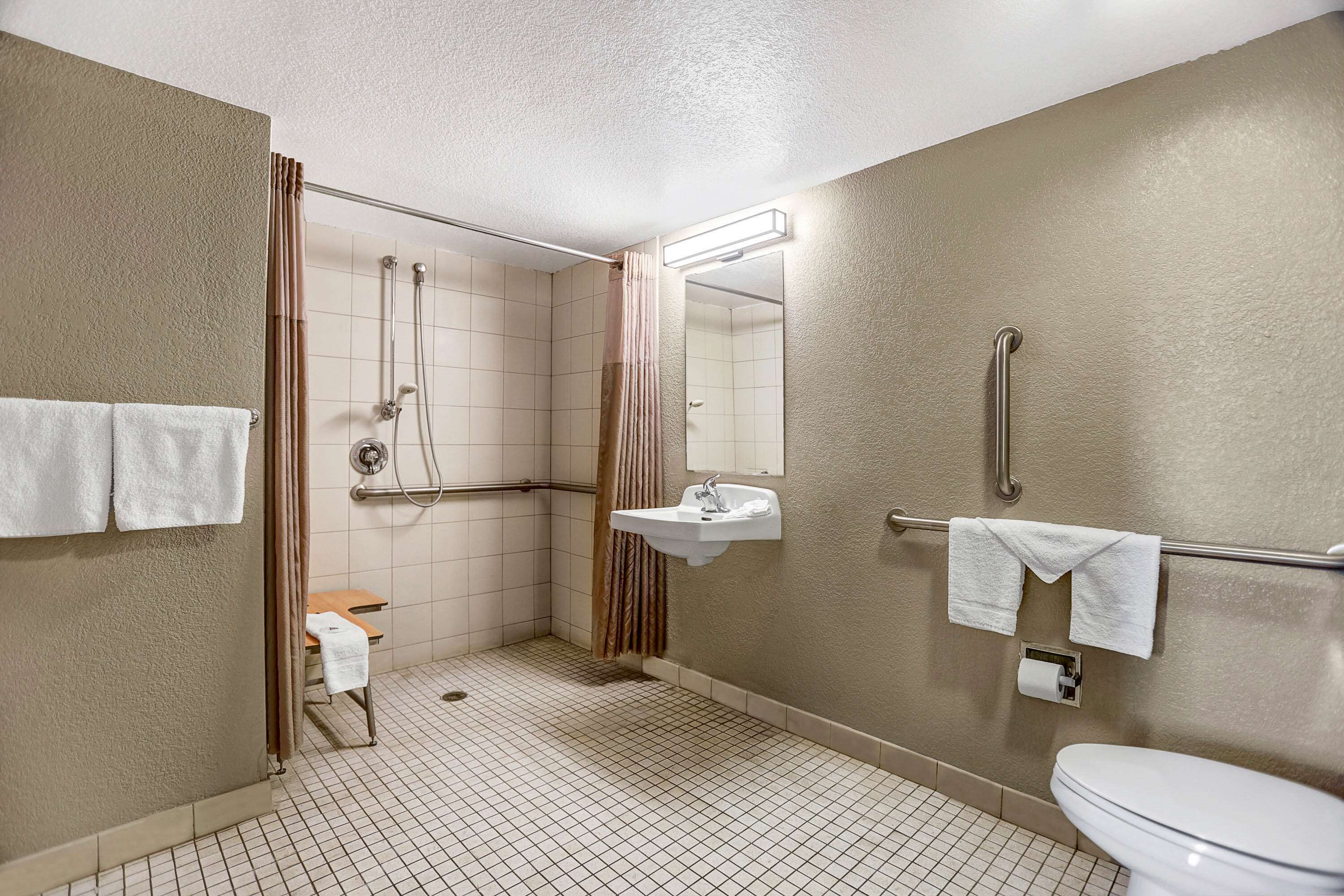 A standard hotel bathroom with a shower, toilet, sink, and handrails.