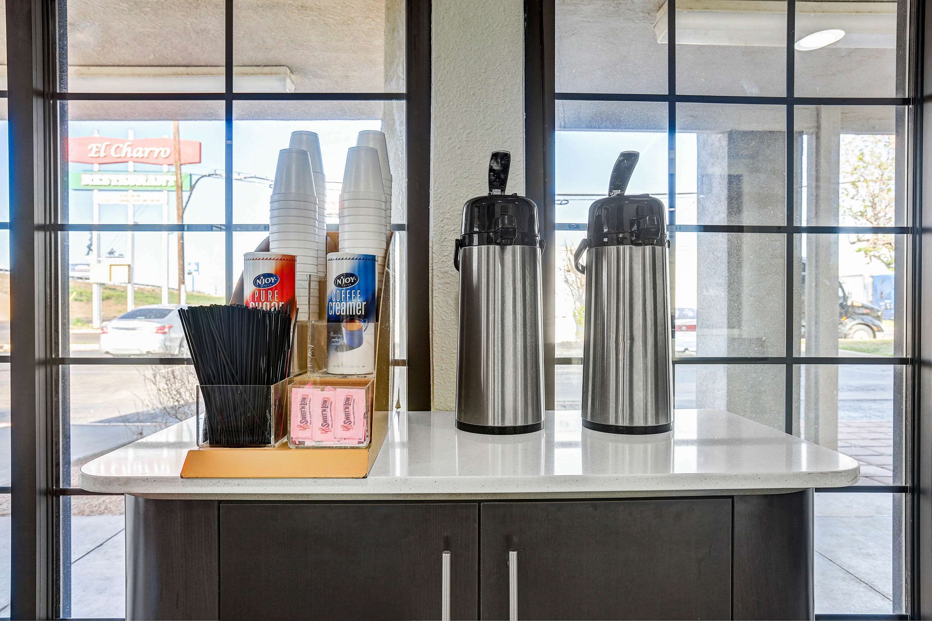 A coffee station with disposable cups, a coffee creamer dispenser, and a thermos.