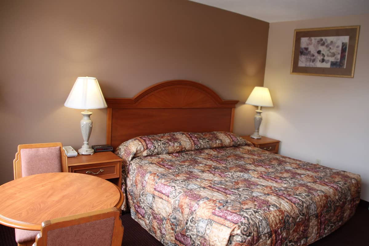Quarto de hotel com cabeceira de madeira, cama com colcha estampada, mesa de madeira redonda, duas luminárias de mesa e um pequeno móvel com gavetas.