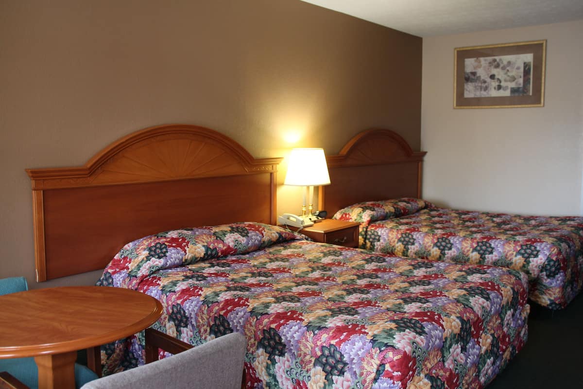 Quarto de hotel com duas camas, cabeceira de madeira, mesa de cabeceira com abajur, mesinha redonda e uma cadeira.