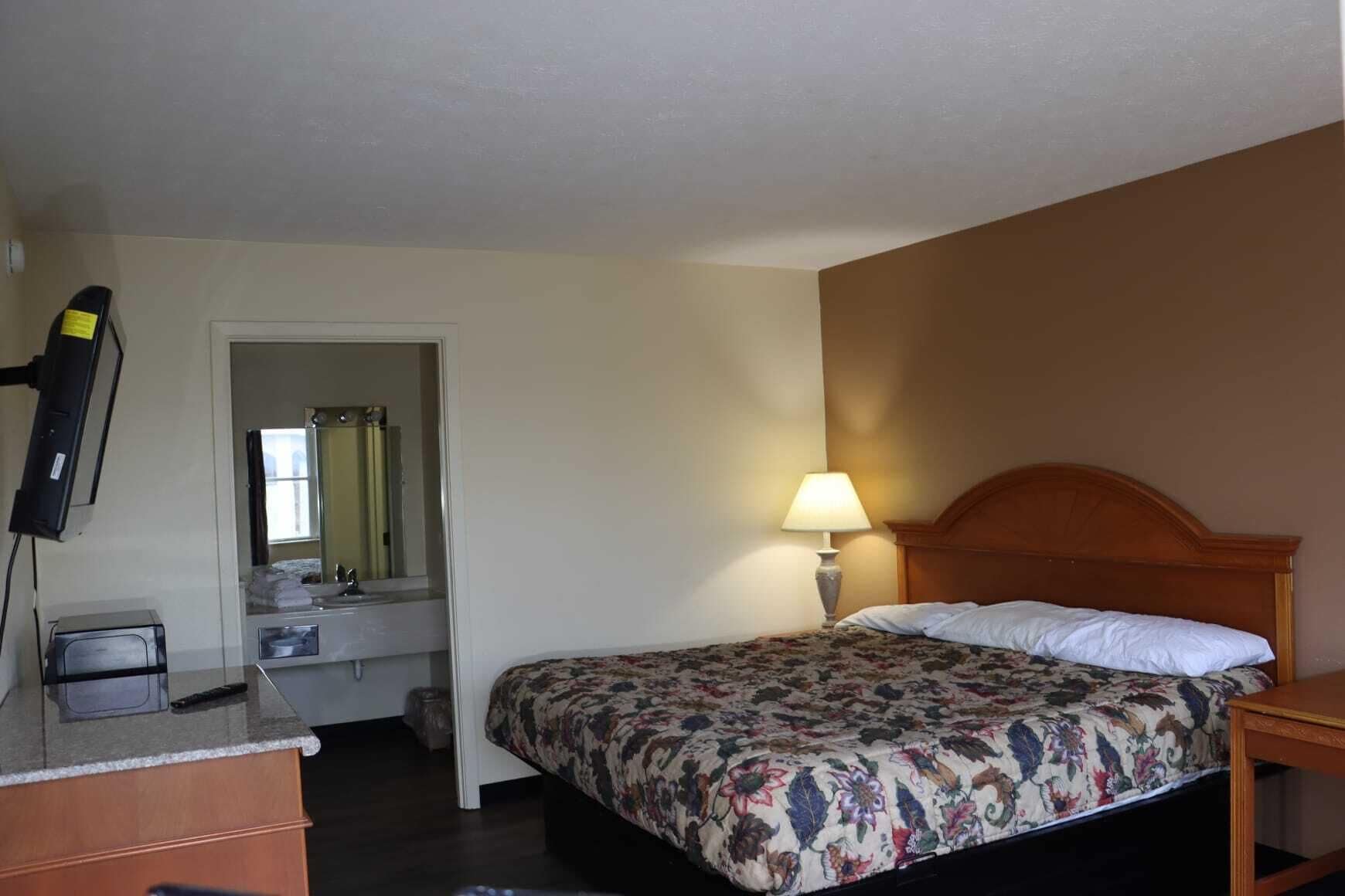 Habitación de hotel con cama, escritorio con lámpara, televisión y baño visible a través de una puerta abierta.