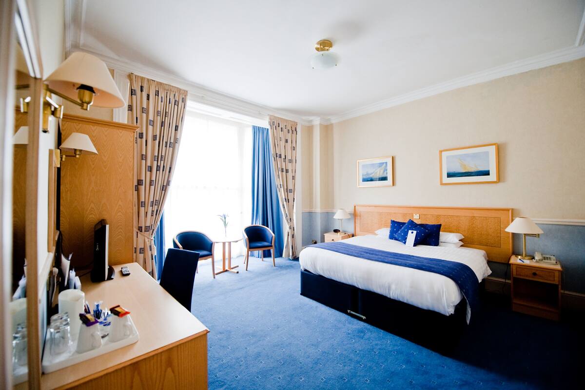 Ein Hotelzimmer mit einem großen Bett, Nachttischen, einem Schreibtisch mit Stuhl und Blick ins Freie.