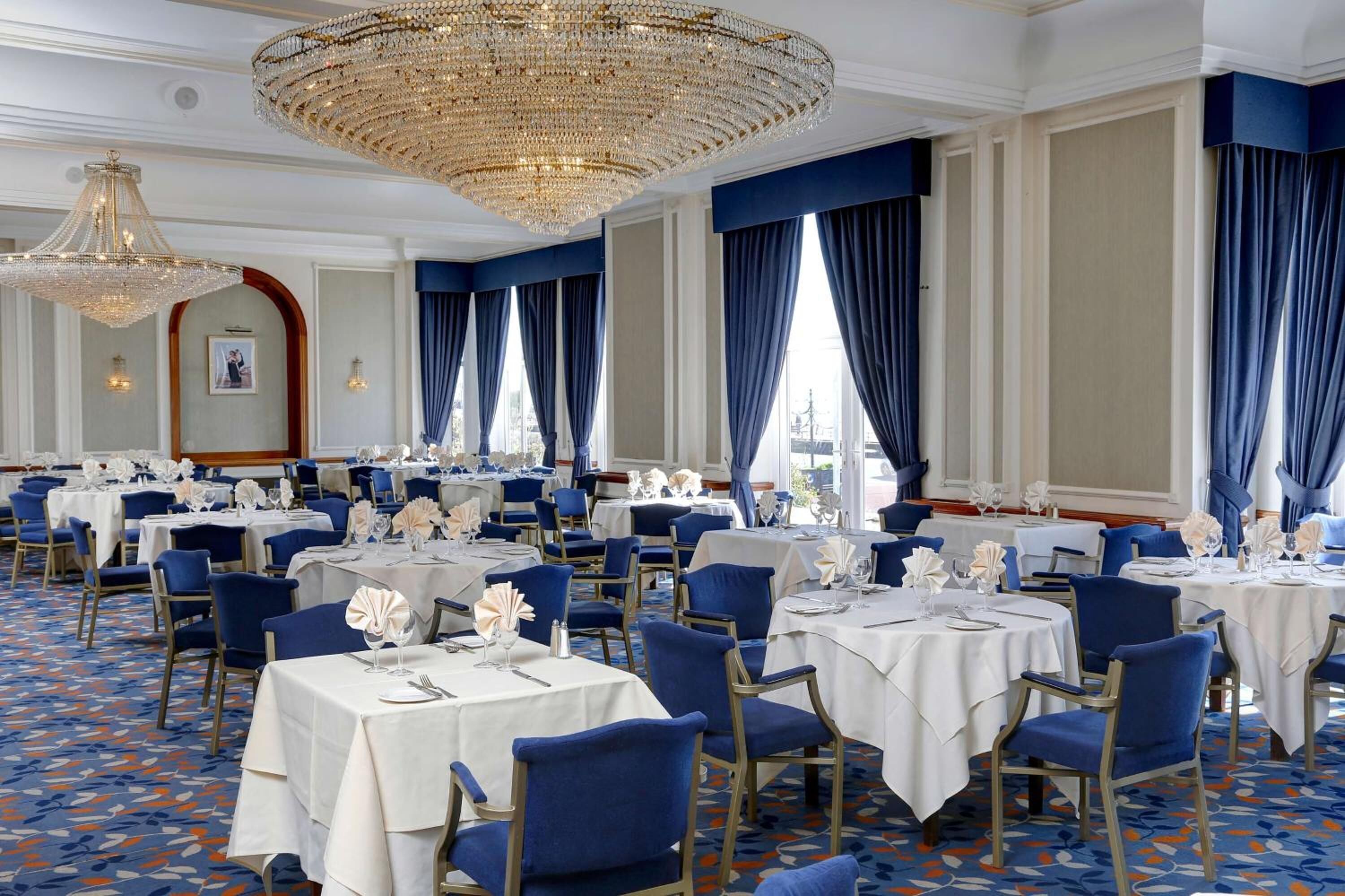 Een klassieke eetzaal met ronde tafels, blauwe stoelen en kroonluchters.