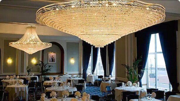 Une salle à manger élégante, avec plusieurs tables dressées pour le service, de grands lustres et une vue sur l’extérieur par les fenêtres.