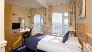 Une chambre d’hôtel comprenant un lit, un bureau avec une télévision, une chaise, une commode et une fenêtre avec des rideaux.