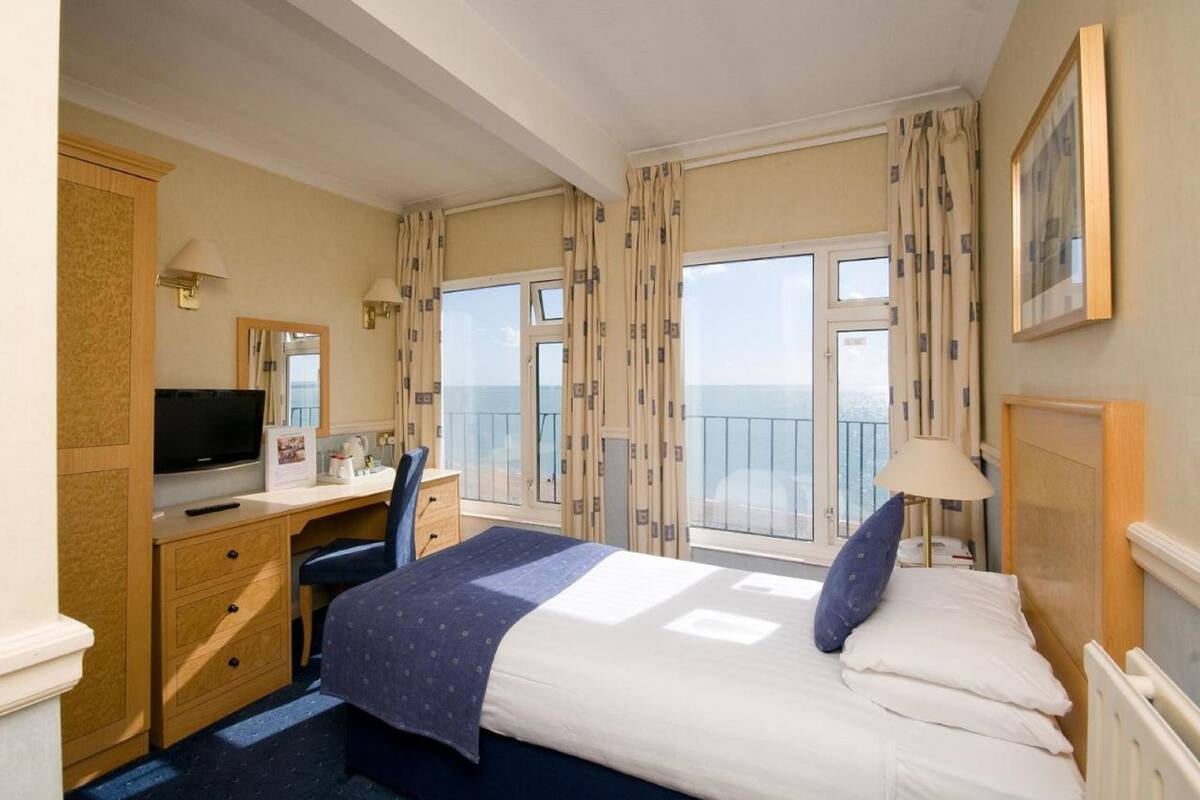 Ein Hotelzimmer mit einem Bett, einem Schreibtisch mit Fernseher, einem Sessel, einer Kommode und einem Fenster mit Vorhängen.