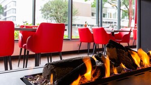 A restaurant interior with a fireplace, red chairs, and a view of the outside. - Bastion Hotel Den Haag Rijswijk (Rijswijk)