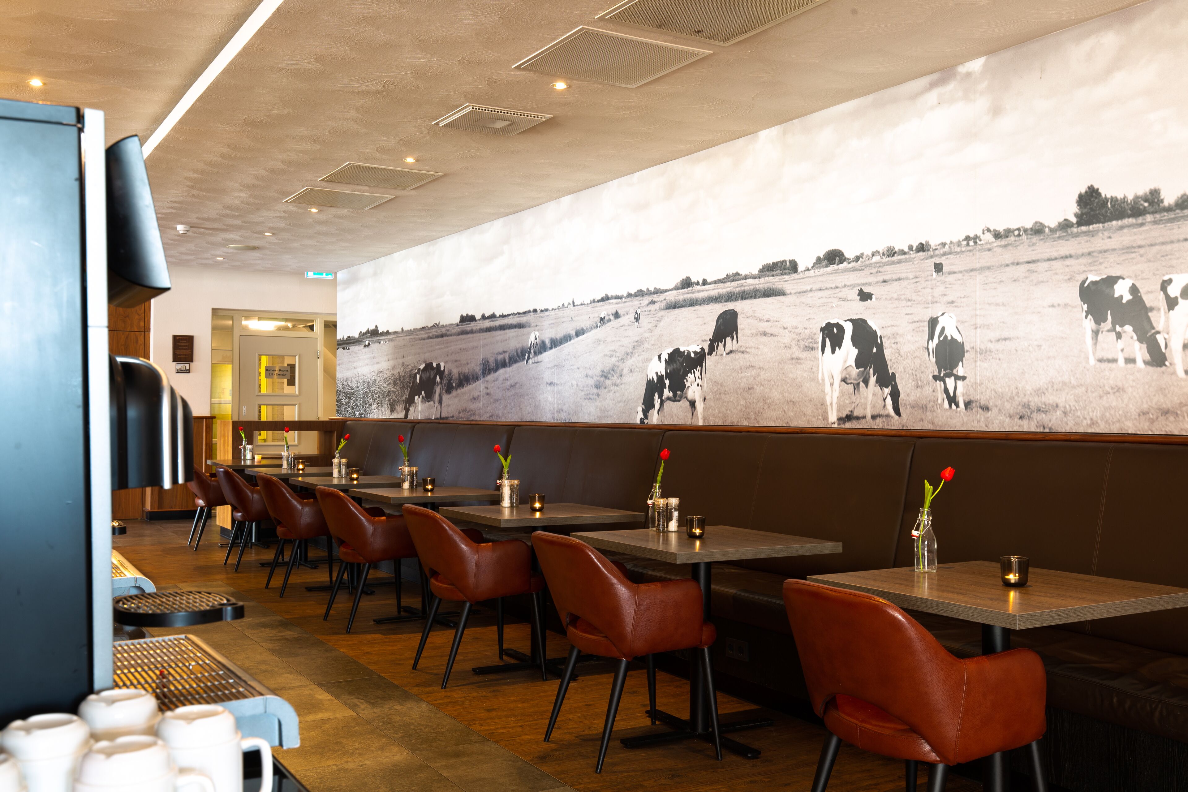 A restaurant with booths, tables, and chairs. There is a wall mural of cows in a field.