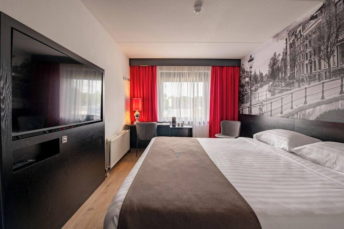 Hotelkamer met een groot bed, een flatscreen televisie, een bureau met stoel en een raam met rode gordijnen.