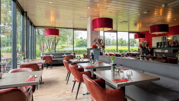 A modern restaurant with wooden flooring, red pendant lights, and a view of greenery outside. - Bastion Hotel Schiphol Hoofddorp (Hoofddorp)