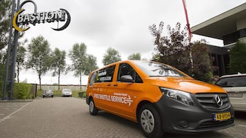 En orange shuttlebus med tekst der siger "Free Shuttle Service" og "Amsterdam Airport", parkeret på en belagt område med træer og en bygning i baggrunden.