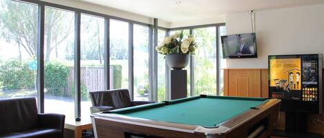 A pool table with a flower vase, a television, and a pool cue rack in a room with large windows and a wooden floor.