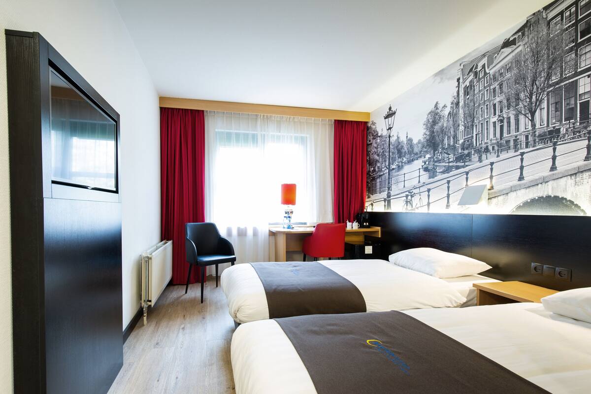 Habitación de hotel con dos camas, un escritorio, una silla y un ventanal con cortinas rojas.