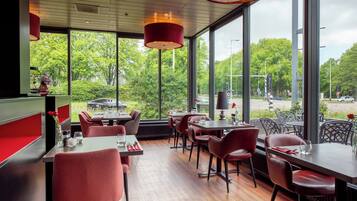 A restaurant with red upholstered chairs and tables, large windows offering a view of trees and a road.