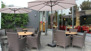 Un espace extérieur aménagé avec des chaises et des tables en osier, protégé par de grands parasols, et un mur de verre en arrière-plan.