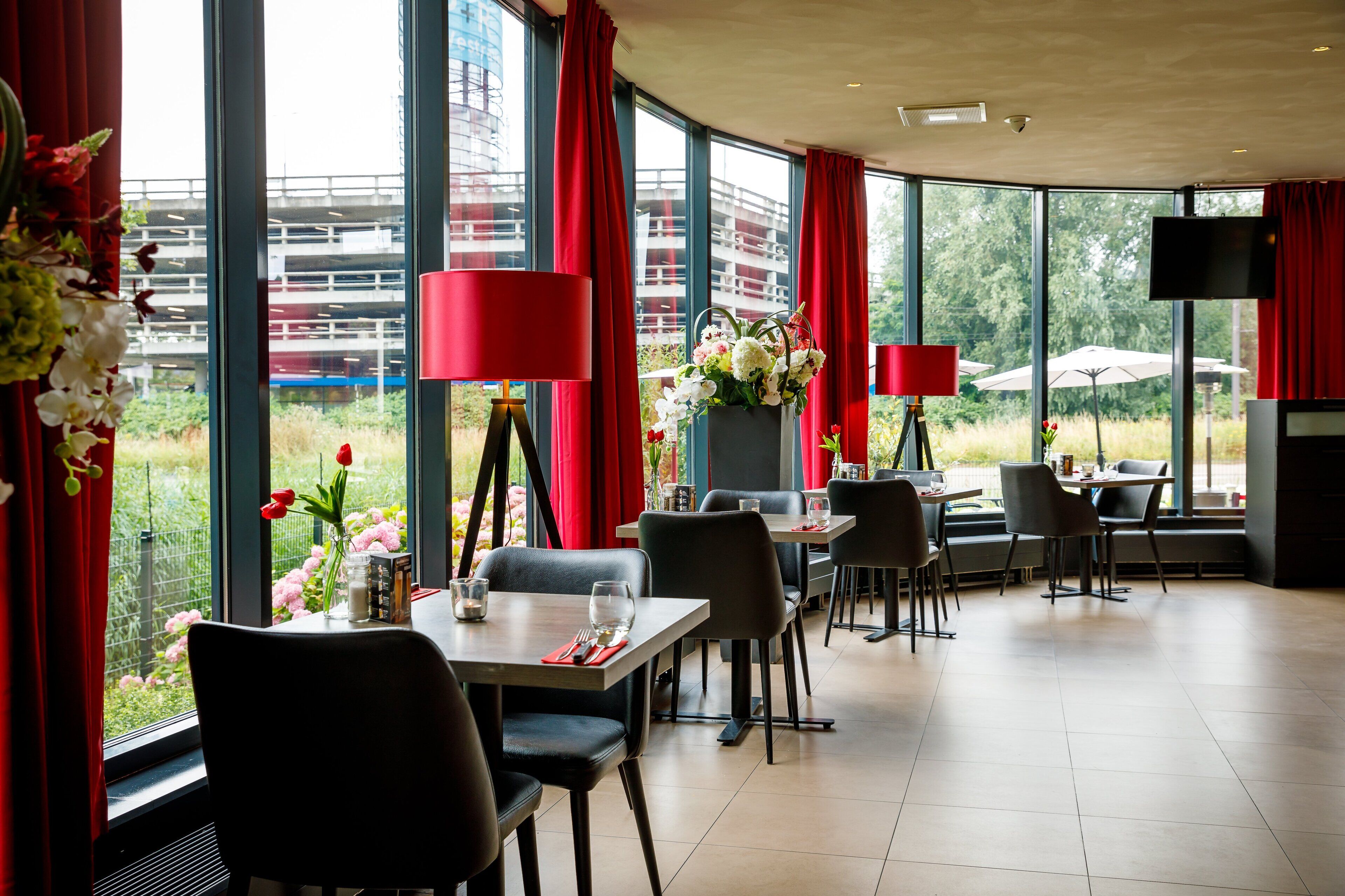 A restaurant with tables and chairs, red curtains, and large windows with a view of buildings and greenery.