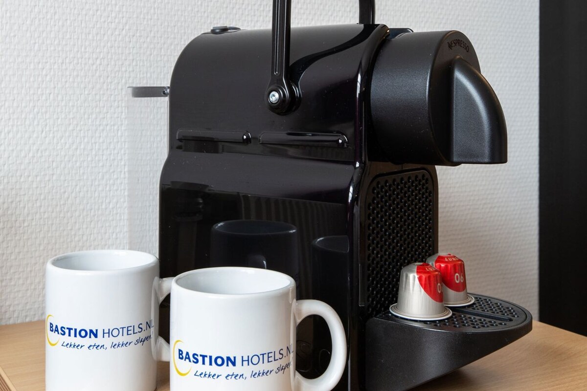 A black espresso machine with two white mugs labeled 'Bastion Hotels.NL' on a wooden surface.