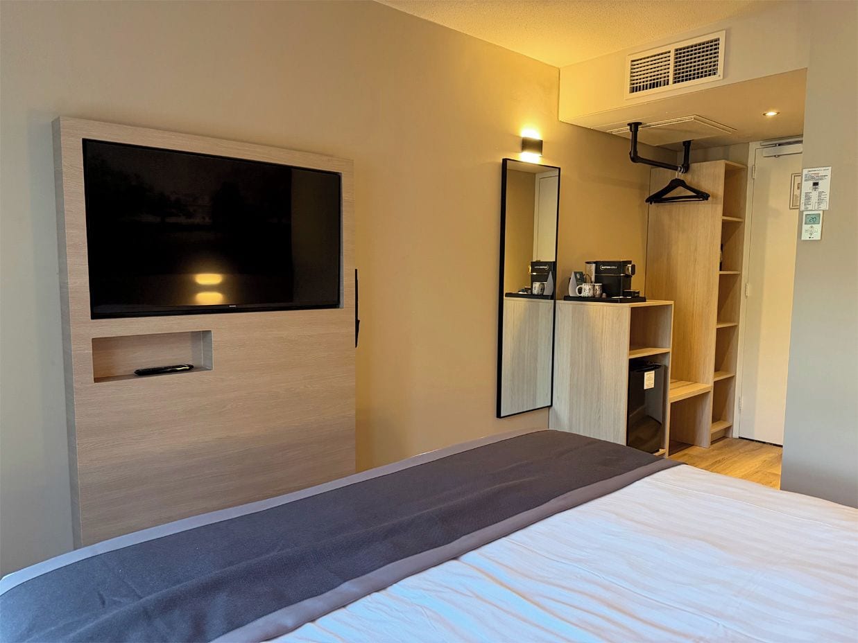 A hotel room with a bed, a TV mounted on the wall, a wardrobe, and a mirror.