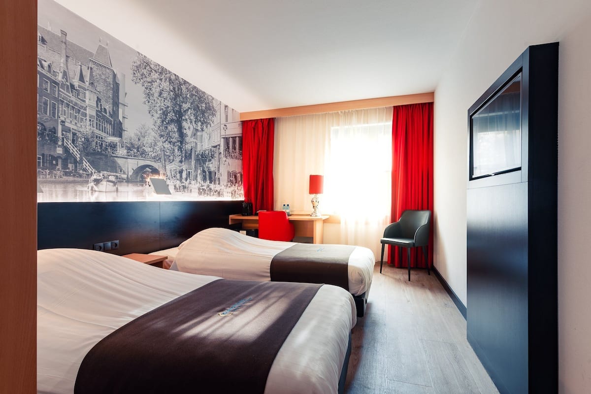 Une chambre d’hôtel avec deux lits, un bureau, une chaise et une grande fenêtre avec des rideaux rouges.