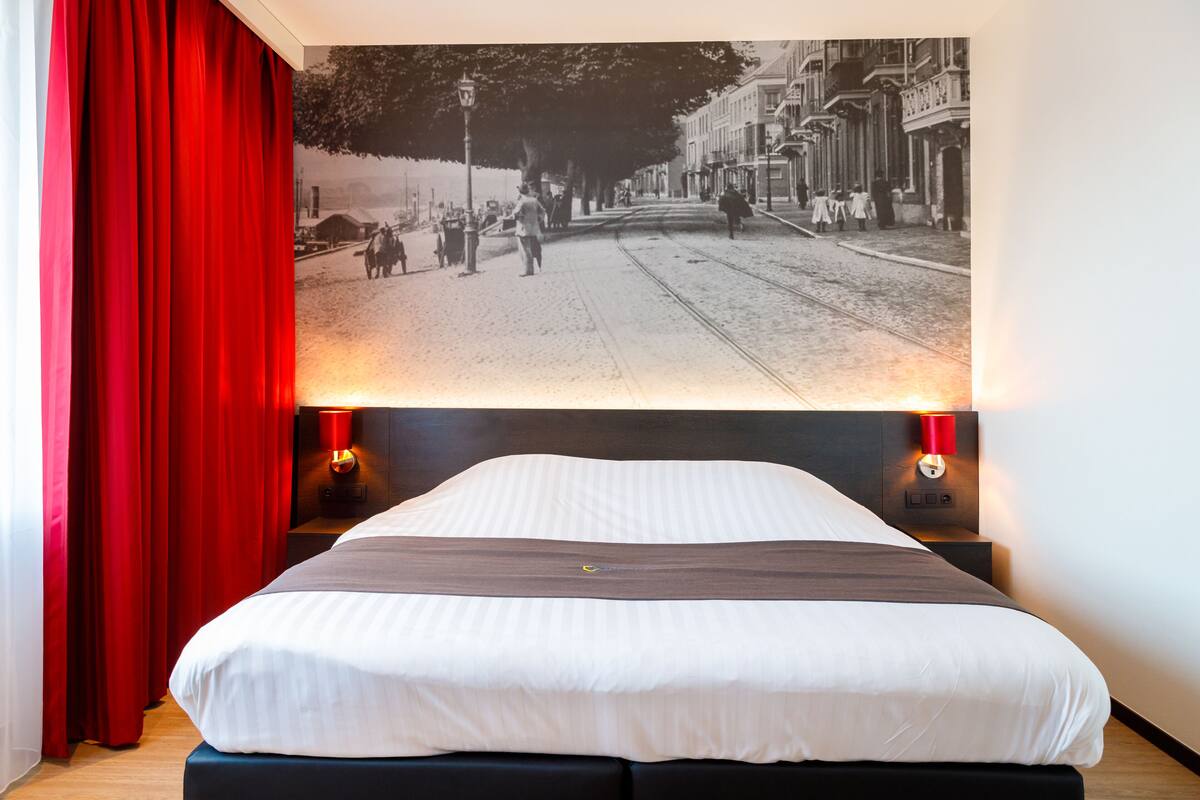 Une chambre d’hôtel avec un grand lit, des rideaux rouges, une tête de lit dotée de lampes murales et une affiche représentant une scène de rue d’époque accrochée au mur.
