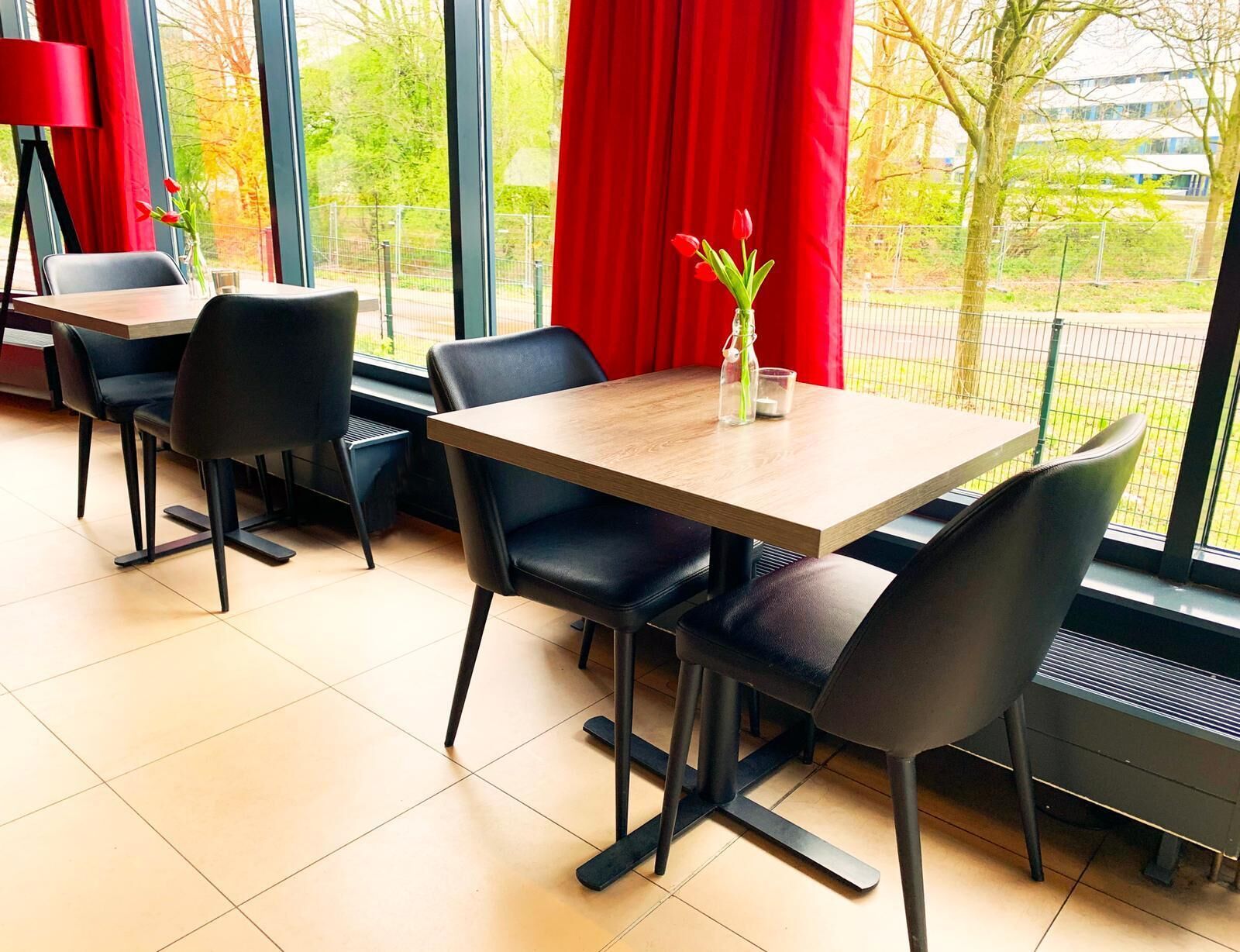 A dining area with a table and chairs, a vase with flowers, and a view of trees and buildings through large windows.