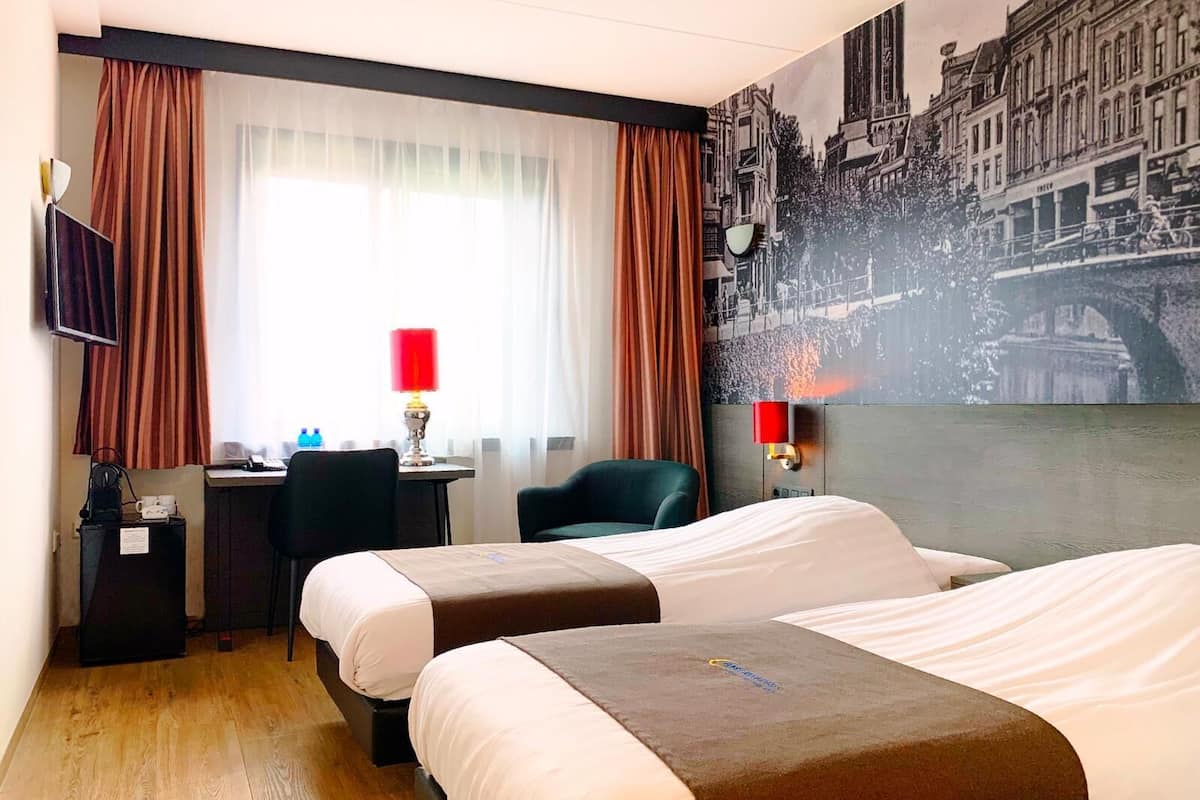Une chambre d’hôtel avec deux lits, un bureau, une chaise, une télévision et une grande fresque murale représentant un paysage urbain.